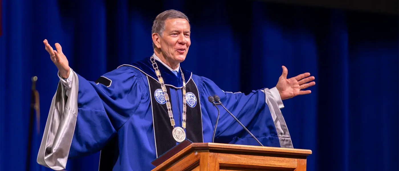 U N E president James D. Herbert stands at podium at graduation in the commencement garb with his arms outstretched gesturing to the crowd